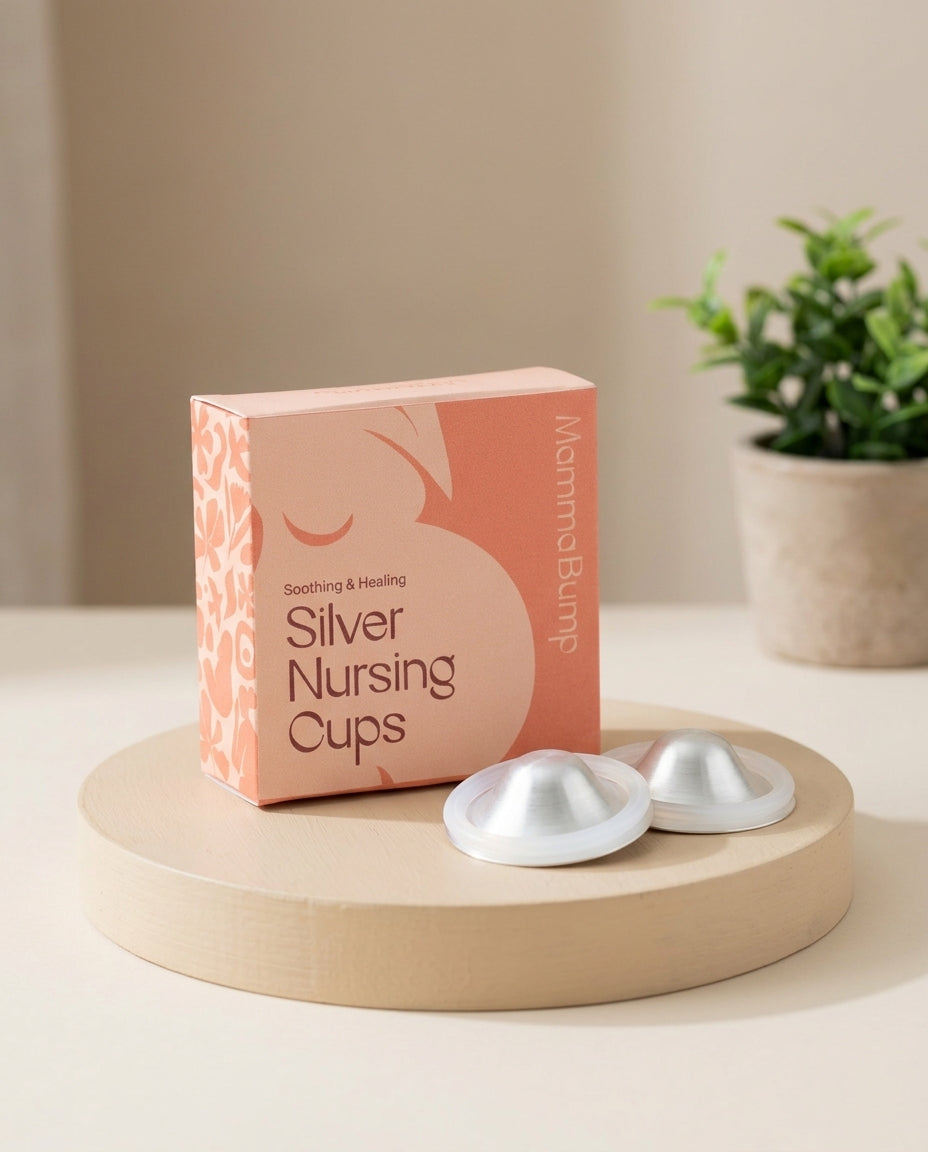 A box of MammaBump Silver Nursing Cups sits on a round wooden pedestal, with two silver silicone cups placed in front. The background is a softly lit indoor setting with a blurred potted plant.