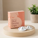 A box of MammaBump Silver Nursing Cups sits on a round wooden pedestal, with two silver silicone cups placed in front. The background is a softly lit indoor setting with a blurred potted plant.