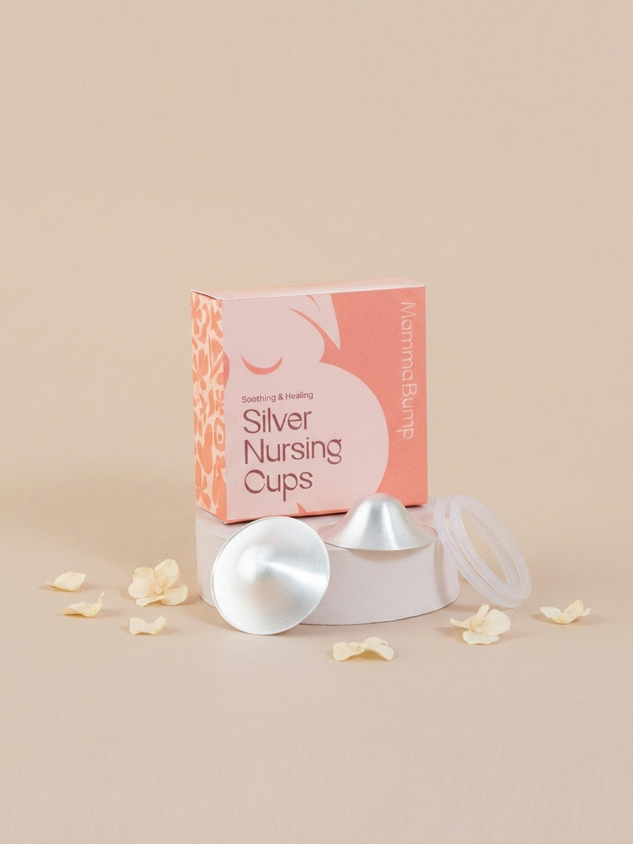 A boxed set of silver nursing cups, silverettes, displayed on a pedestal with two cups and a clear lid in front of a peach and pink box. The box reads 'Breastfeeding Aids Silver Nursing Cups' and 'Alarmed Bump!'.