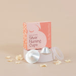 A boxed set of silver nursing cups, silverettes, displayed on a pedestal with two cups and a clear lid in front of a peach and pink box. The box reads 'Breastfeeding Aids Silver Nursing Cups' and 'Alarmed Bump!'.