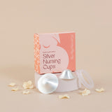 A boxed set of silver nursing cups, silverettes, displayed on a pedestal with two cups and a clear lid in front of a peach and pink box. The box reads 'Breastfeeding Aids Silver Nursing Cups' and 'Alarmed Bump!'.